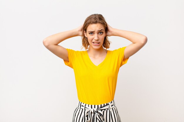 Young blonde woman feeling stressed, worried, anxious or scared, with hands on head, panicking at mistake against flat color wall