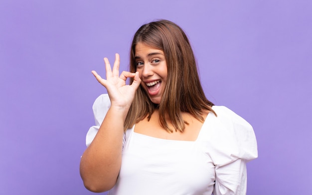 Young blonde woman feeling successful and satisfied, smiling with mouth wide open, making okay sign with hand