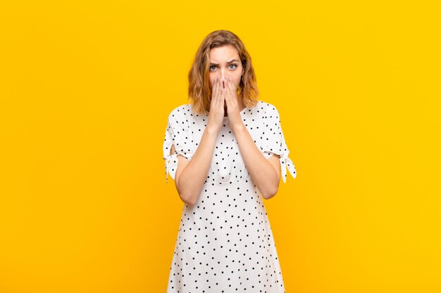 Young blonde woman feeling worried, upset and scared, covering mouth with hands, looking anxious and having messed up over color wall