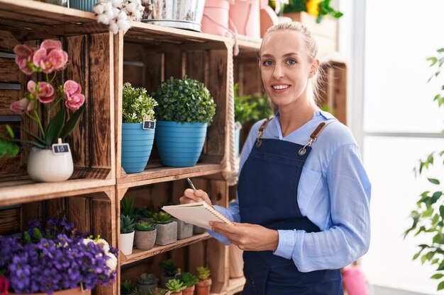 Young blonde woman florist smiling confident writing on notebook at florist