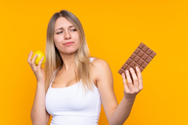 Young blonde woman having doubts while taking a chocolate tablet in one hand and an apple in the other