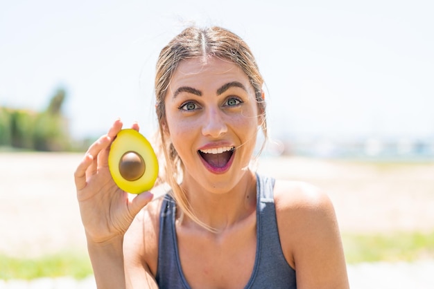 Young blonde woman holding an avocado at outdoors with surprise and shocked facial expression