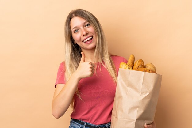 Young blonde woman holding a bag full of breads isolated on beige wall giving a thumbs up gesture