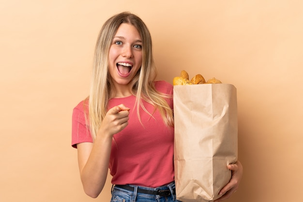 Young blonde woman holding a bag full of breads isolated on beige wall points finger at you