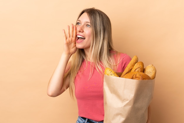 Young blonde woman holding a bag full of breads isolated on beige wall shouting with mouth wide open