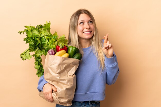Молодая белокурая женщина, держащая сумку, полную овощей по изолированной стене с пересечением пальцев