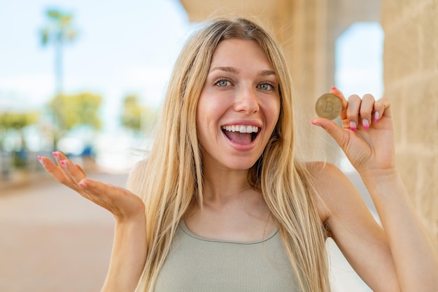 Young blonde woman holding a bitcoin at outdoors with shocked facial expression