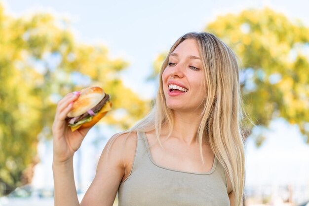 Photo young blonde woman holding a burger at outdoors with happy expression