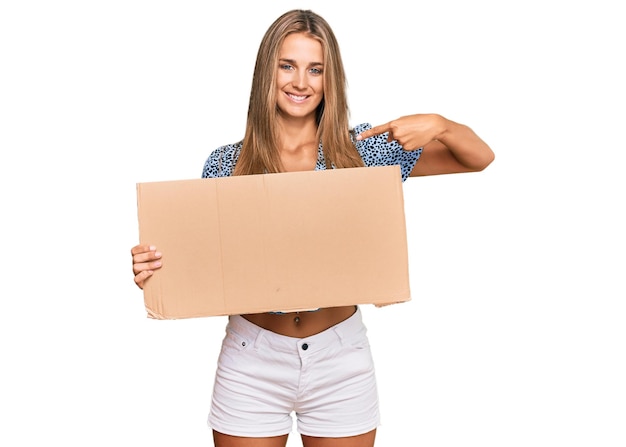 Photo young blonde woman holding cardboard empty banner pointing finger to one self smiling happy and proud