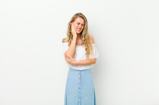 Photo young blonde woman holding cheek and suffering painful toothache, feeling ill, miserable and unhappy, looking for a dentist against white wall