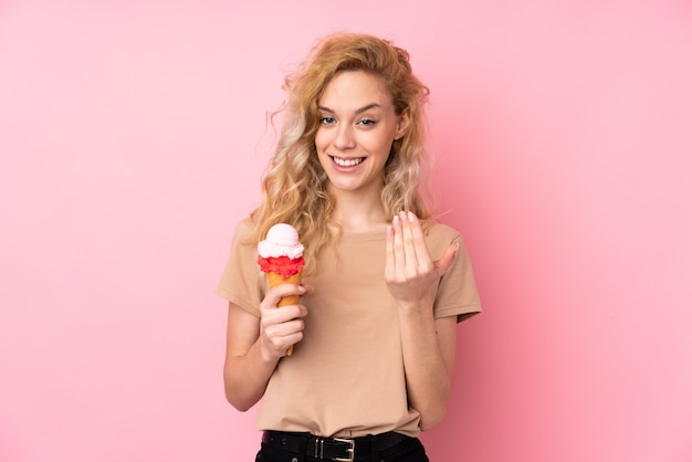 Giovane donna bionda che giudica un gelato della cornetta isolato sulla parete rosa che invita a venire con la mano. felice che tu sia venuto