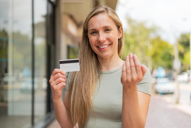 Young blonde woman holding a credit card at outdoors inviting to come with hand Happy that you came