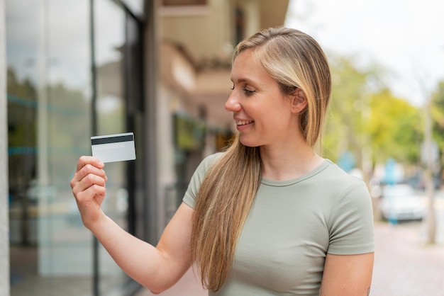 幸せな表情で外でクレジットカードを握っている若い金の女性