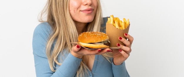 Young blonde woman holding fried chips and cheeseburger over isolated background