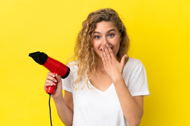 Young blonde woman holding a hairdryer isolated on yellow background happy and smiling covering mouth with hand
