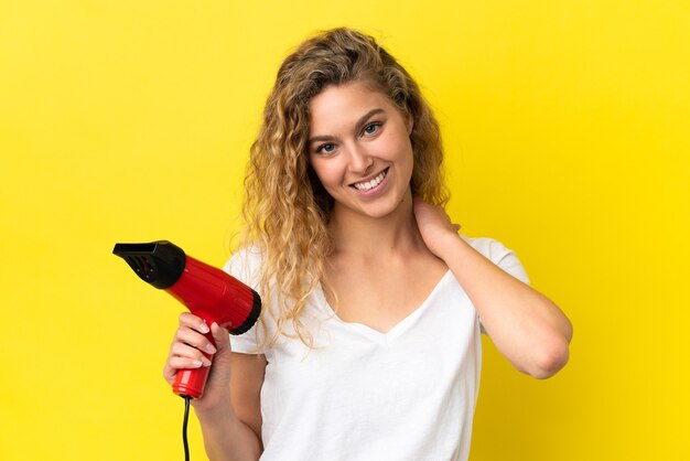 Young blonde woman holding a hairdryer isolated on yellow background laughing