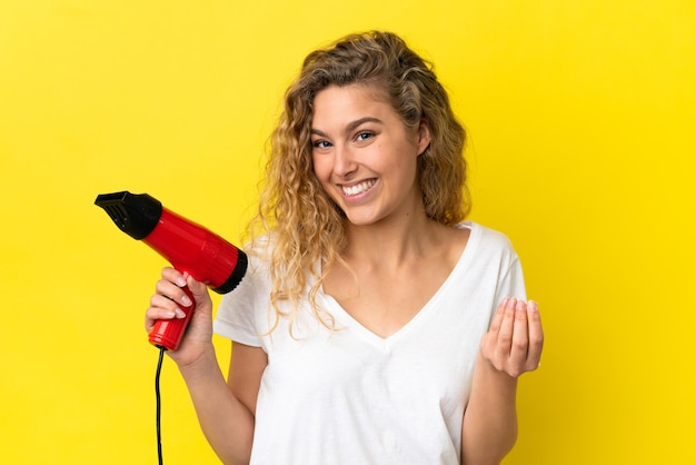 Young blonde woman holding a hairdryer isolated on yellow background making money gesture