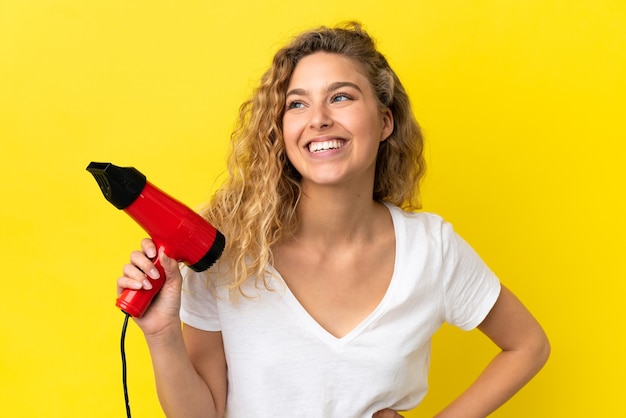 Young blonde woman holding a hairdryer isolated on yellow background posing with arms at hip and smiling