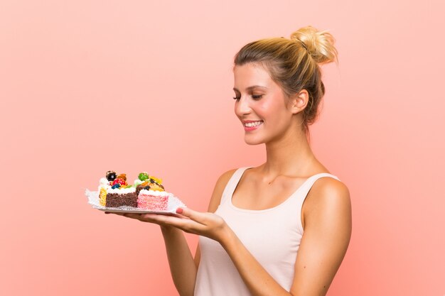 Young blonde woman holding lots of different mini cakes with surprise facial expression