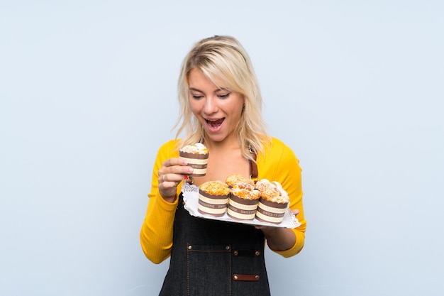 Young blonde woman holding mini cakes and surprised