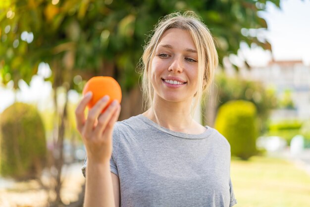 Young blonde woman holding an orange at outdoors with happy expression