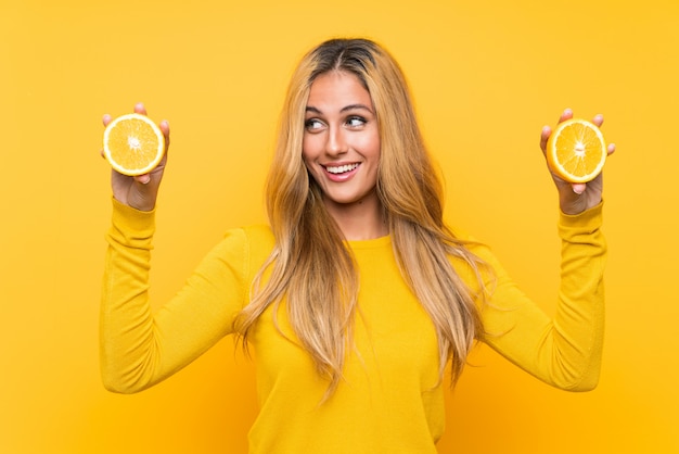 Young blonde woman holding an orange over yellow wall