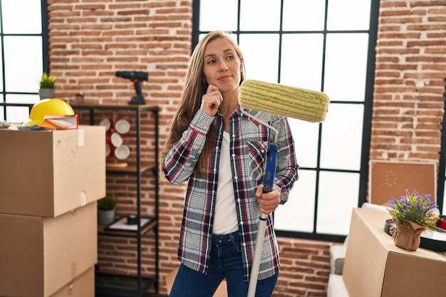 Photo young blonde woman holding paint roller with doubt expression at new home