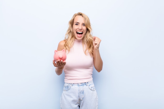 young blonde woman holding a piggy bank feeling excited and happy