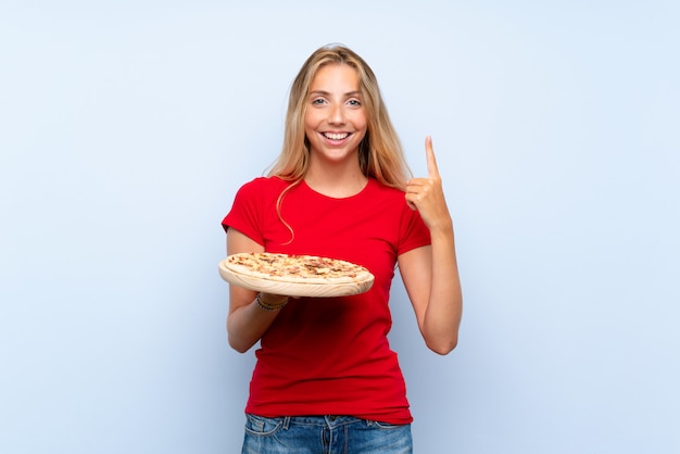 Молодая белокурая женщина держа пиццу над изолированной голубой стеной указывая вверх по отличной идее