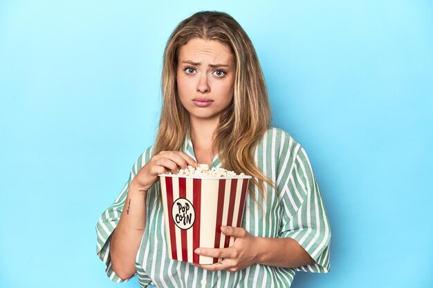 Young blonde woman holding popcorn and a TV remote in a studio