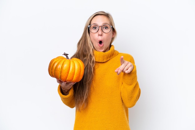 Young blonde woman holding a pumpkin isolated on white background surprised and pointing front