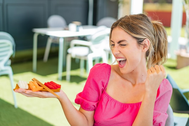 Young blonde woman holding sashimi at outdoors celebrating a victory