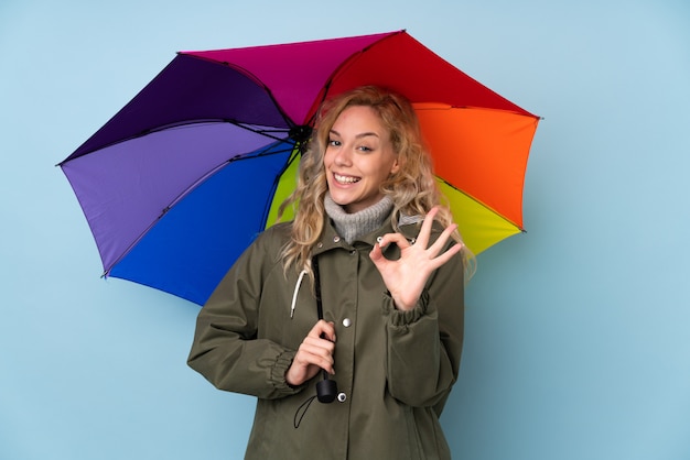 Young blonde woman holding an umbrella on blue showing ok sign with fingers