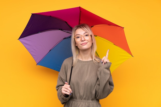 Молодая белокурая женщина держа зонтик над изолированной желтой стеной указывая с указательным пальцем отличная идея