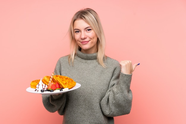 Young blonde woman holding waffles over isolated pink wall pointing to the side to present a product