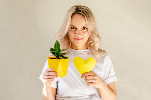 Young blonde woman holding yellow heart and pot plant at home Love for plants Indoor gardening hobby gardening