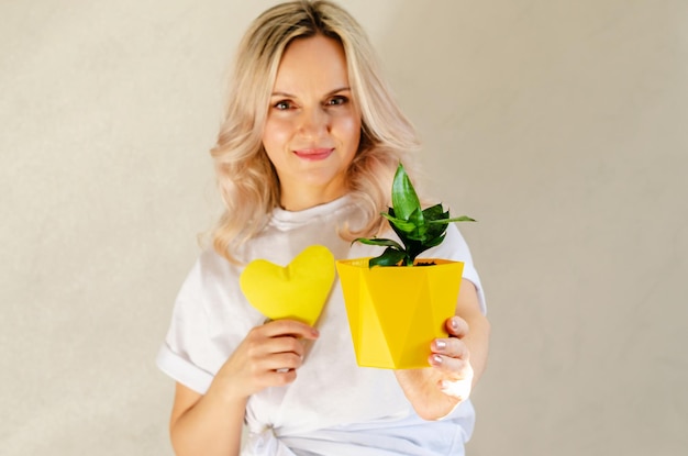 Giovane donna bionda con cuore giallo e pianta in vaso a casa amore per le piante giardinaggio indoor giardinaggio hobby