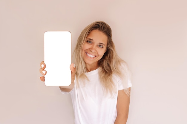 Young blonde woman holds mobile phone in her hand demonstrating white screen for design