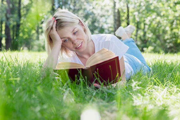 A young blonde woman is lying on the grass in a park on a sunny summer day and is reading a book. Recreation and distance learning in nature.