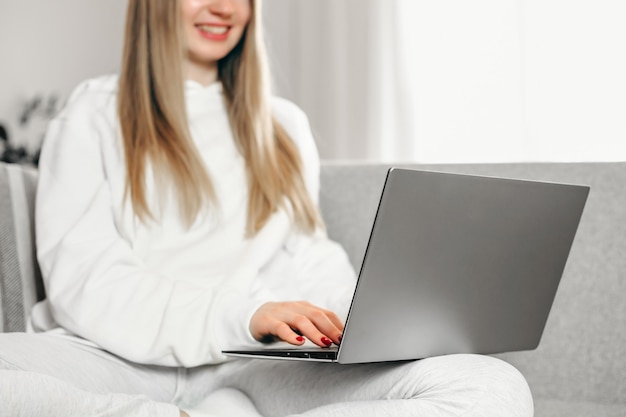 A young blonde woman is sitting on the couch and using her laptop.