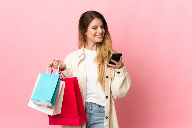 Young blonde woman over isolated background holding shopping bags and writing a message with her cell phone to a friend