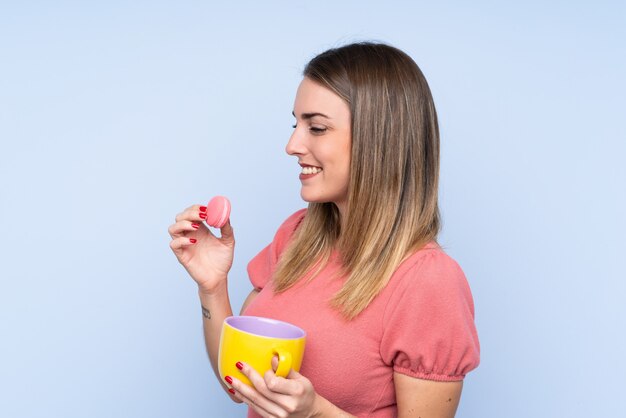 Young blonde woman over isolated blue wall holding colorful French macarons and a cup of milk