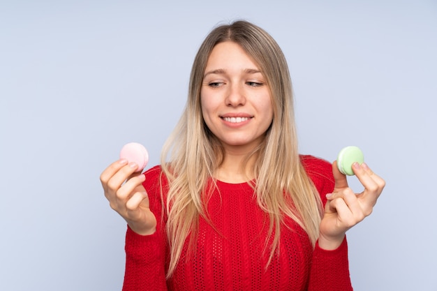 Young blonde woman over isolated blue wall holding colorful French macarons and looking it