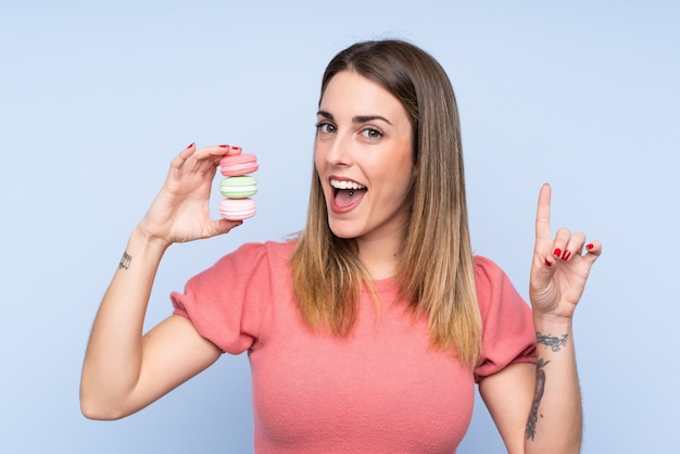 Young blonde woman over isolated blue wall holding colorful French macarons and pointing up a great idea