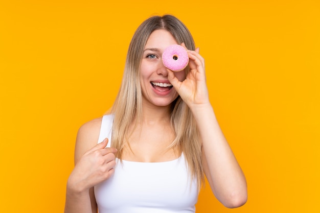 Young blonde woman over isolated blue wall holding a donut and happy