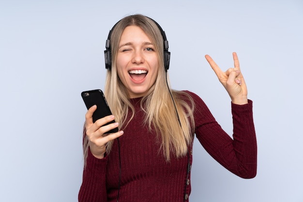 Young blonde woman over isolated blue wall listening music with a mobile making rock gesture