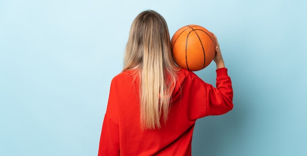 Young blonde woman isolated on blue wall playing basketball in back position