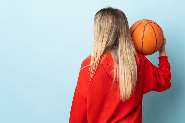 Young blonde woman isolated on blue wall playing basketball in back position