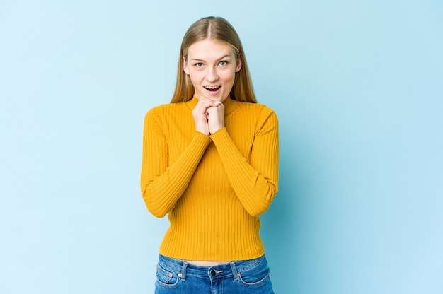 Young blonde woman isolated on blue wall praying for luck, amazed and opening mouth looking to front.