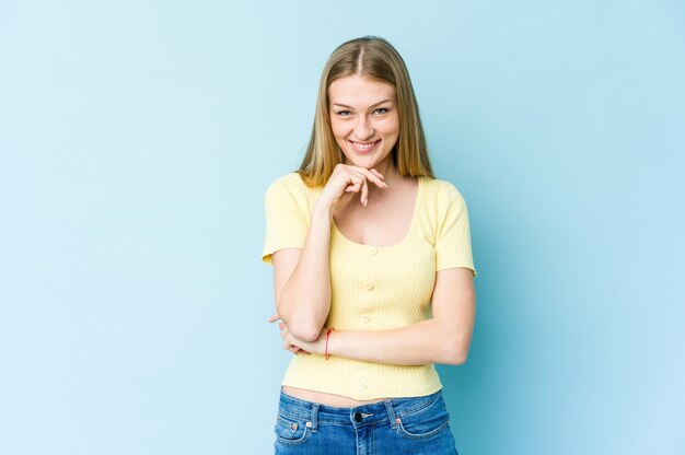 Young blonde woman isolated on blue wall smiling happy and confident, touching chin with hand
